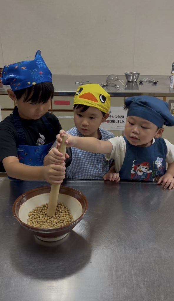 すり鉢を囲み、3人でいっしょにすりこぎを持つ子どもたち